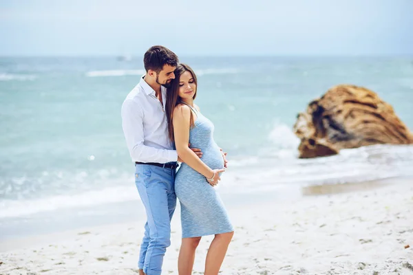 Courageous and handsome man in a white shirt and blue trousers  hugs her beautiful pregnant wife in a blue dress against the background of the rocks and the sea. — Stock Photo, Image