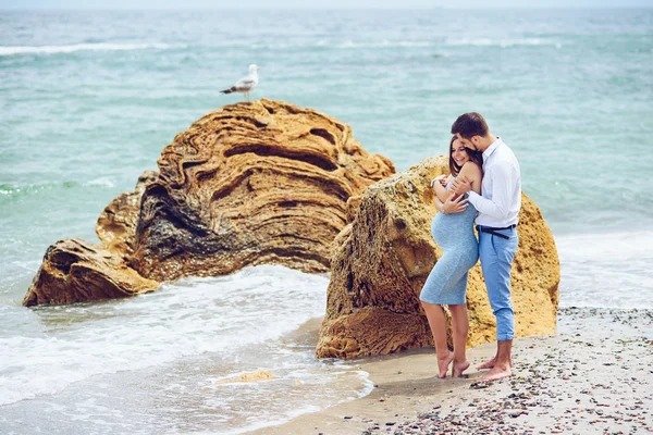 Una bella donna incinta in un abito blu e il suo marito coraggioso e bello in camicia e pantaloni blu appoggiati l'uno all'altro sullo sfondo della roccia con gabbiano e il mare . — Foto Stock