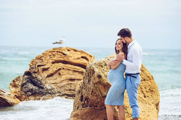 Una bella donna incinta in un abito blu e il suo marito coraggioso e bello in camicia e pantaloni blu appoggiati l'uno all'altro sullo sfondo delle rocce con gabbiano e il mare . — Foto Stock