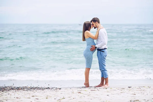 Una bella donna incinta in un abito blu baciare al naso il suo marito coraggioso e bello in camicia e pantaloni blu sullo sfondo del mare . — Foto Stock