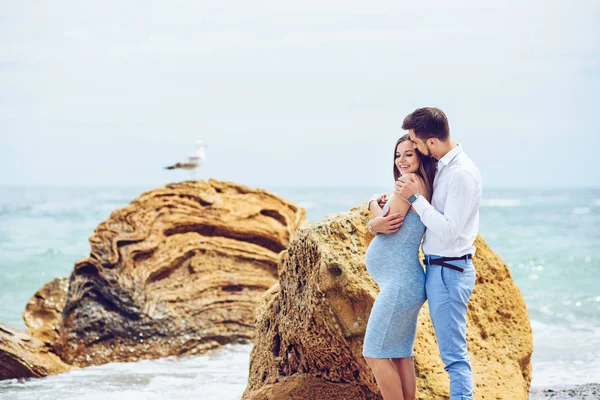 青いドレスの美しい妊婦とシャツと青いズボンで彼女の勇気とハンサムな夫は、かもめと海と岩の背景に互いに寄りかかって. — ストック写真