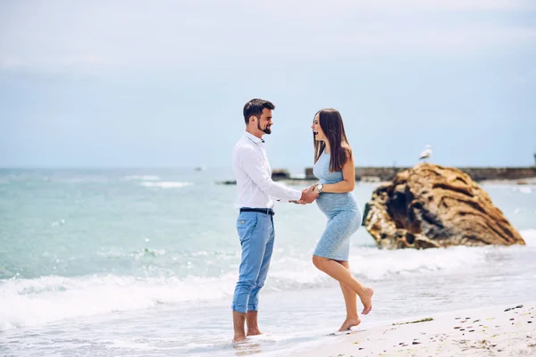 Una bella donna incinta sorridente in un abito blu e il suo marito coraggioso e bello in camicia e pantaloni blu appoggiati l'uno all'altro sullo sfondo delle rocce e del mare . — Foto Stock