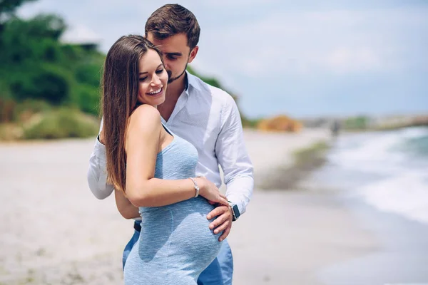 Hombre valiente y guapo en una camisa blanca y pantalones azules abraza y besa en una mejilla a su hermosa esposa embarazada en un vestido azul sobre el fondo del mar . — Foto de Stock