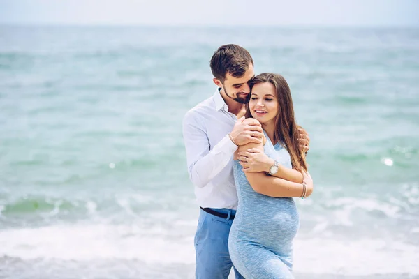 Coraggioso e bello uomo in una camicia bianca e pantaloni blu abbraccia la sua bella moglie incinta in un abito blu sullo sfondo del mare . — Foto Stock