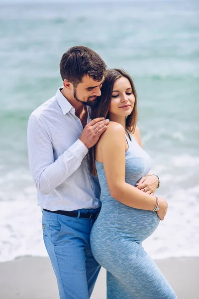 Courageous and handsome man in a white shirt and blue trousers  hugs her beautiful pregnant wife in a blue dress against the background of the sea. — Stock Photo, Image
