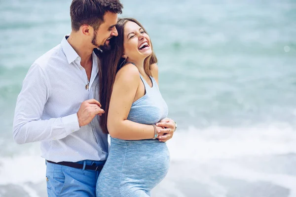 Hombre valiente y guapo en una camisa blanca y pantalones azules abraza a su hermosa esposa embarazada sonriente en un vestido azul contra el fondo del mar . — Foto de Stock