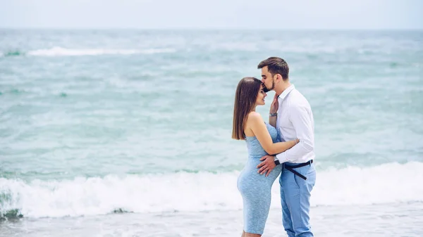 Homem corajoso e bonito em uma camisa branca e calças azuis beijando na testa sua bela esposa grávida em um vestido azul contra o fundo das rochas e do mar . — Fotografia de Stock