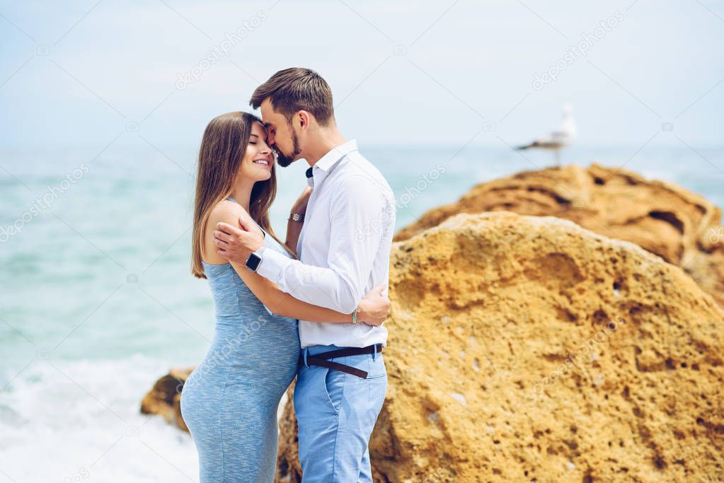 A beautiful pregnant woman in a blue dress and her courageous and handsome husband in a shirt and blue trousers leaned to each other against the background of the rock and the sea.