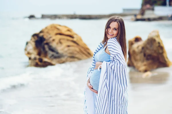 Güzel bir hamile kadın sıkı elbise onun göbekle dokunmadan sahilde beyaz çizgili ekose kaplı bir mavi sevgi ve kayalar ve deniz arka plan üzerinde bakım.