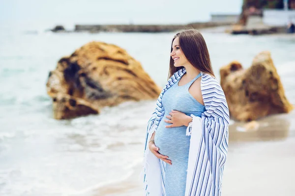 Une belle femme enceinte en robe bleue serrée est recouverte d'un plaid rayé blanc sur la plage touchant son ventre avec amour et soin sur le fond des rochers et de la mer . — Photo