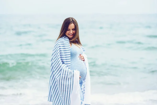 Une belle femme enceinte en robe bleue serrée est recouverte d'un plaid rayé blanc sur la plage qui touche son ventre avec amour et soin. Promenade au bord de la mer par une journée ensoleillée . — Photo