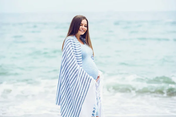 A beautiful pregnant woman in a blue tight dress is covered with a white striped plaid on the beach touching her belly with love and care. Walking on the seashore in sunny day. — Stock Photo, Image