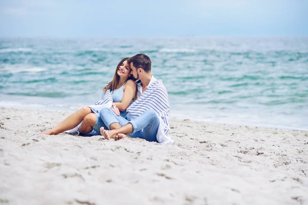 Bella donna incinta e il suo uomo rispettabile appoggiato l'uno contro l'altro e coperto con un plaid a strisce bianche seduto sulla riva del mare. Bella giovane coppia . — Foto Stock