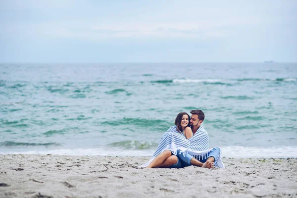 Bella donna incinta e il suo uomo rispettabile appoggiato l'uno contro l'altro e coperto con un plaid a strisce bianche seduto sulla riva del mare. Bella giovane coppia . — Foto Stock