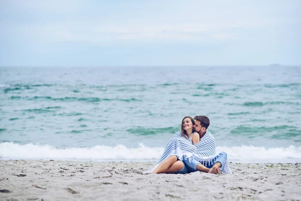 Mulher grávida bonita e seu homem respeitável encostados uns contra os outros e coberto com uma xadrez listrado branco sentado na praia. Adorável jovem casal . — Fotografia de Stock