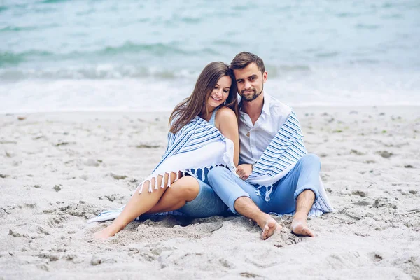 Szép terhes nő és tiszteletre méltó ember hajlik egymás ellen, és borított egy fehér csíkos, kockás ül a tengerparton. Szép fiatal pár. — Stock Fotó