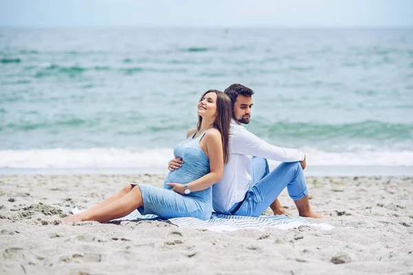 Bella donna incinta e il suo uomo rispettabile appoggiato l'uno contro l'altro e seduto sulla riva del mare. Bella giovane coppia . — Foto Stock