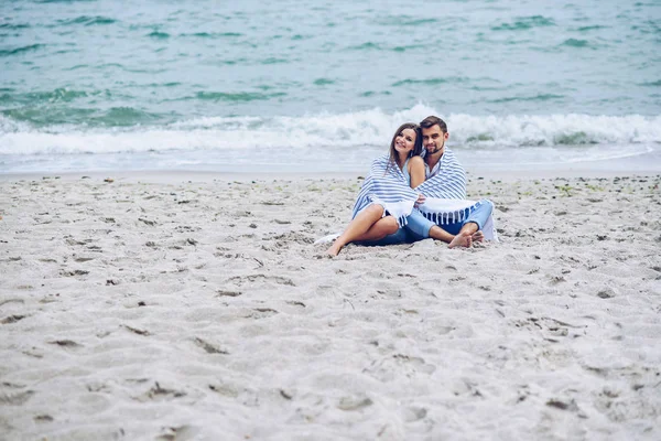 美しい妊娠中の女性と彼女の立派な男互いにもたれて、白で覆われたストライプ プラッドは海辺に座っています。素敵な若いカップル. — ストック写真