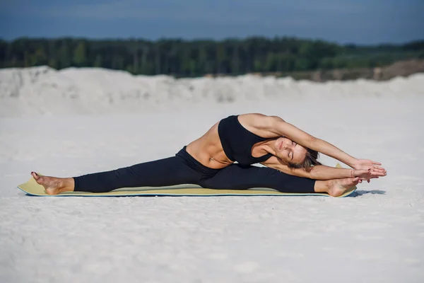 Time for yoga. Attractive and healthy young woman doing exercises on the beach at sunrise or sunset. Fitness girl doing stretching on yoga mat.