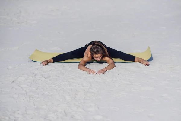 Adorable sporty girl doing twine on yoga mat at the beach. Morning warm up. Fitness sport yoga and healthy lifestyle concept.