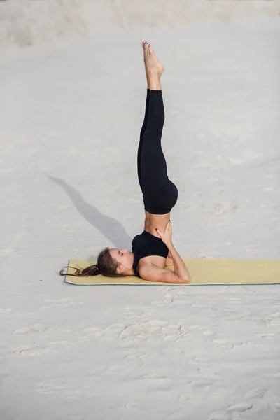Time for yoga. Attractive and healthy young woman doing exercises on the beach at sunrise or sunset. Fitness girl doing stretching on yoga mat.