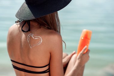 Omzuna arkadan görünüm güneş kremi kalp şeklinde yaz şapkalı güzel bir kız. Kumsalda güneşlenirken kadın. Güneşten korunma.