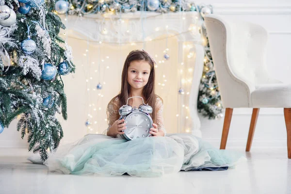 Menina bonito com grande relógio nas mãos se senta perto da árvore de Natal dentro de casa . — Fotografia de Stock