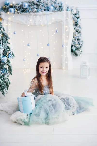 Petite fille mignonne avec boîte cadeau dans une belle robe se trouve près de l'arbre de Noël sur fond de cheminée . — Photo