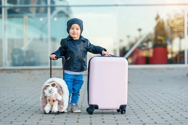 Cute little uśmiechnięta dziewczyna w stylowe ubrania casual z zabawną puszystą walizką na lotnisku machając ręką. — Zdjęcie stockowe