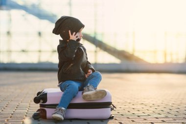 Küçük gezgin kız pembe bir bavula oturur ve gün batımına karşı havaalanı yakınında akıllı telefondan konuşur..