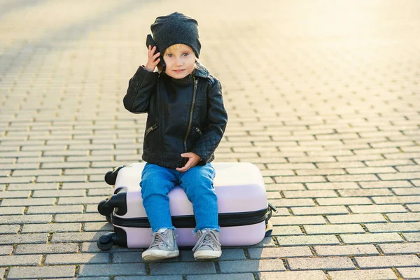 Piękna mała podróżniczka siedzi na różowej walizce i rozmawia przez telefon komórkowy w pobliżu lotniska na tle zachodu słońca. — Zdjęcie stockowe