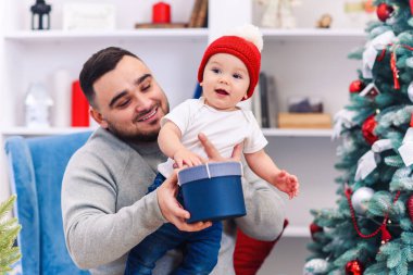 Genç baba, elinde komik bir bebekle rahat koltukta oturuyor ve Noel 'i kutlamak için muhteşem dekore edilmiş odasında ona hediye kutusu veriyor..