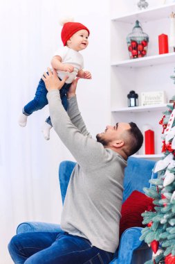 Yakışıklı baba, süslü Noel ağacının yanında komik bebeğini havaya fırlatıyor..