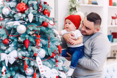 Erkek ve şirin bebek yeni yılı kutlamaya hazırlar. Mutlu baba Noel ağacının yanında küçük oğluyla oynuyor..