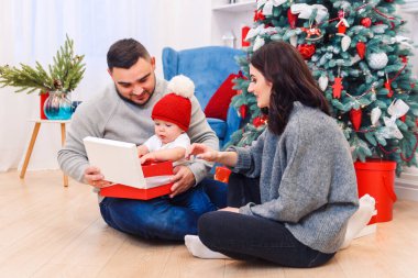 Genç ebeveynler yeni doğan çocuklarına ilk Noel hediyesini açmaya yardım ederler. Küçük çocuk ilk Noel hediyesini aldı..