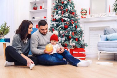 Genç ebeveynler yeni doğan çocuklarına ilk Noel hediyesini açmaya yardım ederler. Küçük çocuk ilk Noel hediyesini aldı..