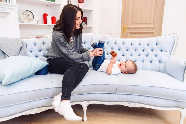 Bonito criança encontra-se no sofá azul e feliz jovem mamãe amassar as pernas e fazê-lo feliz . — Fotografia de Stock