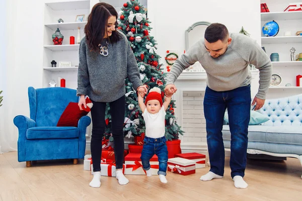 Feliz jovem mãe e pai segurando seu doce bebê pelas mãos e levantando-os no alto da sala decorada para celebrar o Natal — Fotografia de Stock