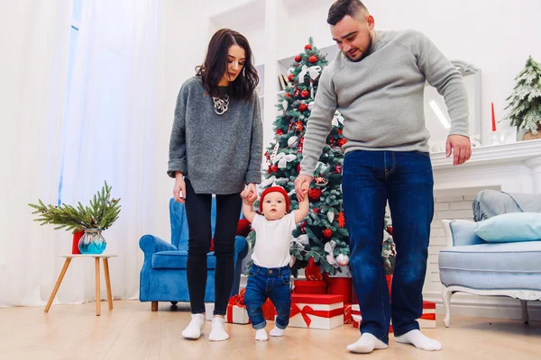 Mãe e pai segurando seu bebê bonito e ensinando-o a andar no meio do quarto decorado de Natal . — Fotografia de Stock