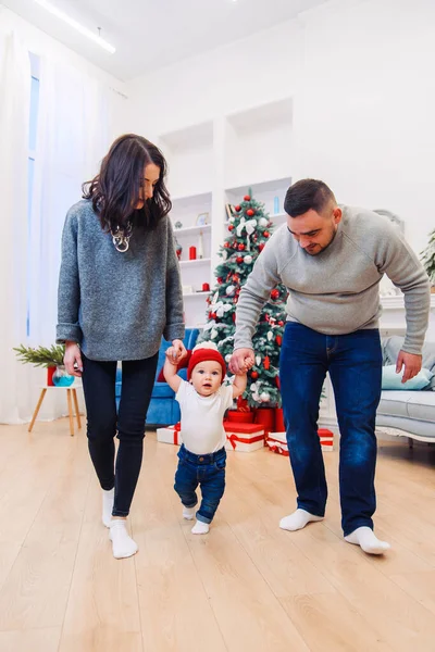 Mãe e pai segurando seu bebê bonito por mãos e ensinando-o a andar no meio da sala de Natal decorado . — Fotografia de Stock