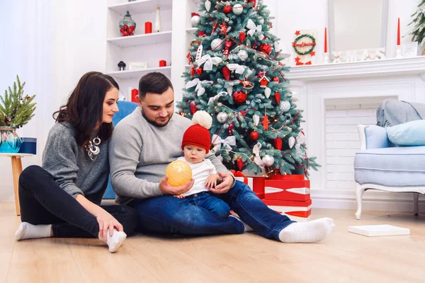 Os pais jovens ajudam a desempacotar o primeiro presente de Natal para seu filho recém-nascido. Menino recebe seu primeiro presente de Natal . — Fotografia de Stock