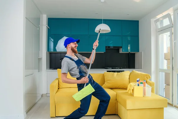 Funny picture of male cleaning worker in special clothes playing with a mop as a guitar in the kitchen