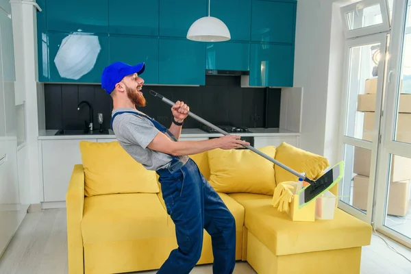 Funny man cleaning worker in special blue clothes singing with a mop as a microphone like a rockstar in the stylish cuisine. Having fun at workplace. — Stock Photo, Image