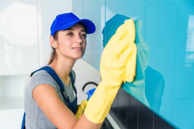 İş elbiseleri içinde sevimli genç bir kadın ve modern yüksek teknoloji mutfağındaki mobilyaları temizleyen koruyucu lastik sarı eldivenler..