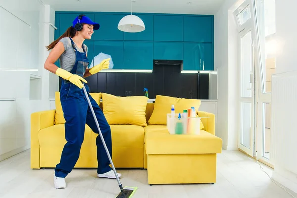 Femme caucasienne comme un nettoyant professionnel dans le plancher de nettoyage casque avec balai et écoute de la musique à la maison . — Photo