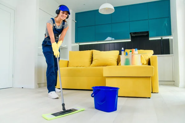 Beautiful young female professional cleaner in special uniform with headphones washing the floor with mop and listens to music at apartment.