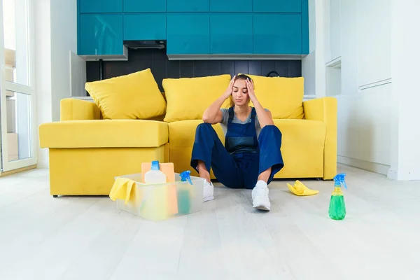 ソファの近くに座って料理の上で床を洗って疲れを感じる清掃サービスの若い女性労働者. — ストック写真