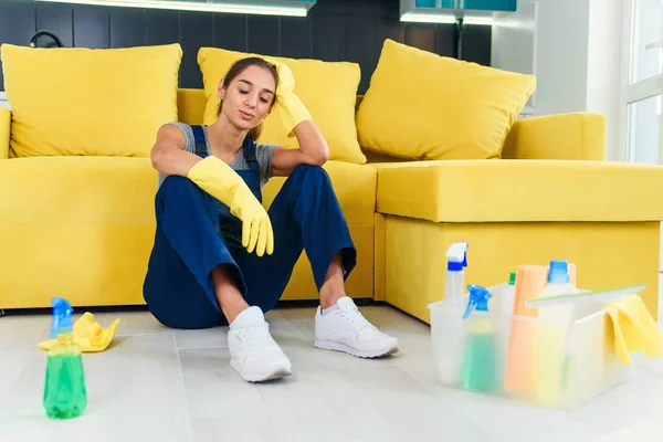 Young female worker of the cleaning service sitting near the couch and feeling tired after washing floor on cuisine.