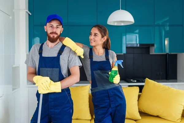 Glimlachende knappe man en aantrekkelijke jonge vrouw als professionele schoonmakers blijven samen in de hedendaagse keuken met wasmiddelen. — Stockfoto