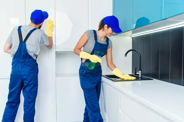 Привлекательная молодая пара мужчин и женщин в синей униформе чистят мебель салфетками и спреем в удобной кухне — стоковое фото
